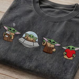 May include: Dark gray t-shirt featuring embroidered images of a green alien character. The alien is depicted holding a coffee cup, in a spaceship, waving, and holding a red heart. The shirt has a crew neck.