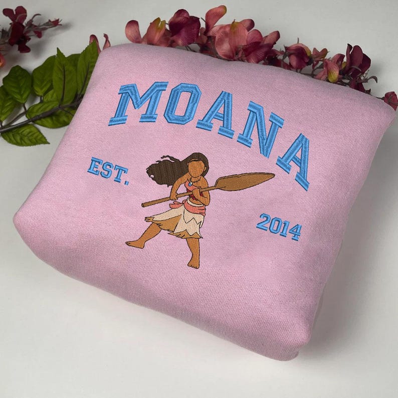 K&ouml;nnte beinhalten: Rosafarbener Pullover mit einem blauen gestickten Motiv von Moana, die ein Paddel h&auml;lt. Der Text "Moana Est. 2014" ist ebenfalls auf dem Pullover gestickt.
