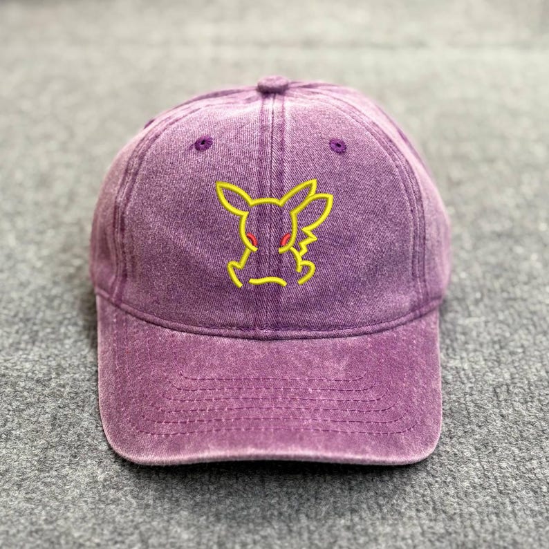 May include: Purple baseball cap with a yellow embroidered cartoon character design. The cap has a curved brim and a fabric strap closure. The hat is made of a soft, washed cotton material.