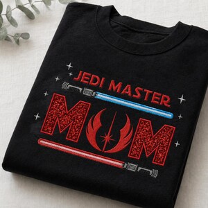 May include: Black t-shirt with the embroidered text "JEDI MASTER MOM" in red and white. The design includes a blue and red lightsaber, and a red emblem. The shirt is folded on a white surface.