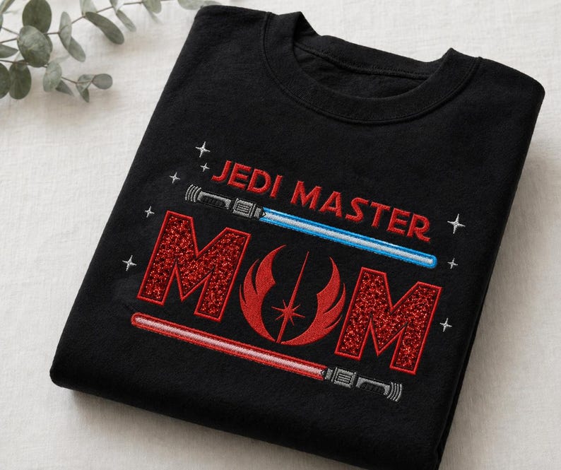 May include: Black t-shirt with the embroidered text "JEDI MASTER MOM" in red and white. The design includes a blue and red lightsaber, and a red emblem. The shirt is folded on a white surface.