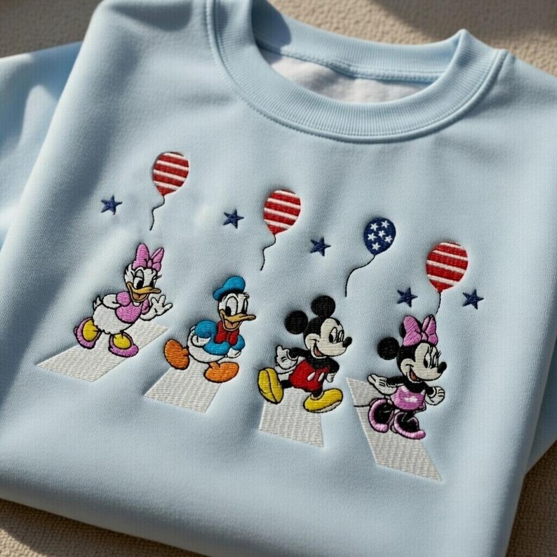 Puede incluir: Sudadera azul claro con bordados de los personajes de Disney Daisy Duck, Donald Duck, Mickey Mouse y Minnie Mouse cruzando una calle. Tambi&eacute;n hay bordados de globos y estrellas rojos, blancos y azules.
