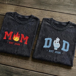 May include: Two dark gray t-shirts with embroidered designs. One shirt says "MOM" in red with a flame graphic and "EST 2026". The other says "DAD" in blue with a cartoon character and "EST 2026".