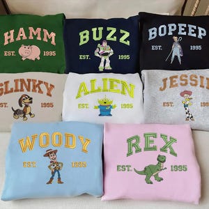 May include: A collection of sweatshirts in various colors, each embroidered with a character name from the movie "Toy Story" in arched lettering. The sweatshirts feature characters like Hamm, Buzz, Bo Peep, Slinky, Alien, Jessie, Woody, and Rex, with "EST. 1995" below.