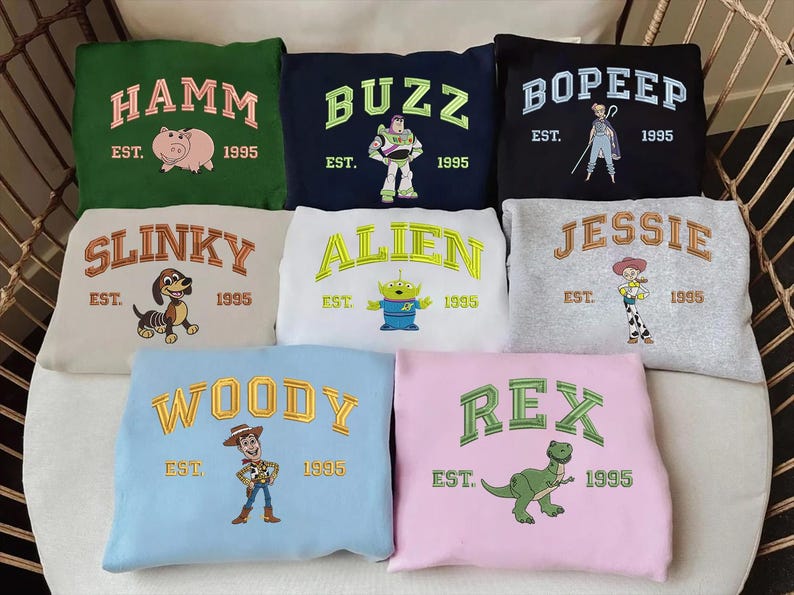 May include: A collection of sweatshirts in various colors, each embroidered with a character name from the movie "Toy Story" in arched lettering. The sweatshirts feature characters like Hamm, Buzz, Bo Peep, Slinky, Alien, Jessie, Woody, and Rex, with "EST. 1995" below.