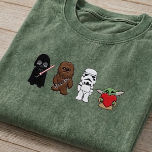 May include: Green t-shirt with embroidered Star Wars characters: Darth Vader, Chewbacca, a Stormtrooper, and Grogu holding a heart. The shirt has a crew neck and a faded look.