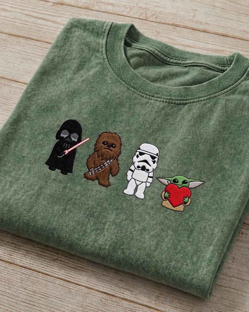 May include: Green t-shirt with embroidered Star Wars characters: Darth Vader, Chewbacca, a Stormtrooper, and Grogu holding a heart. The shirt has a crew neck and a faded look.