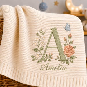 Op de afbeelding: Een crèmekleurige gebreide deken met een geborduurd ontwerp van de letter A in saliegroen, omringd door bloemmotieven in roze en groen, een kleine blauwe vlinder en de naam Amelia geborduurd in cursief schrift onder de letter.