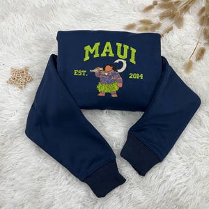 K&ouml;nnte beinhalten: Ein marineblauer Sweatshirt mit einem gestickten Design eines Comic-Charakters, der einen Haken h&auml;lt. Das Design ist in Gr&uuml;n und Gelb gehalten und enth&auml;lt den Text "MAUI EST. 2014".
