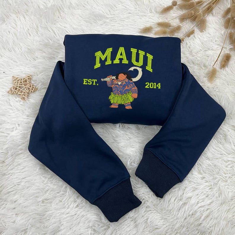 K&ouml;nnte beinhalten: Ein marineblauer Sweatshirt mit einem gestickten Design eines Comic-Charakters, der einen Haken h&auml;lt. Das Design ist in Gr&uuml;n und Gelb gehalten und enth&auml;lt den Text "MAUI EST. 2014".