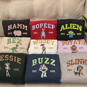 May include: A collection of embroidered sweatshirts featuring characters from the movie Toy Story. Each sweatshirt displays a character's name, the year 1995, and a corresponding image. Colors include black, red, green, white, and gray.