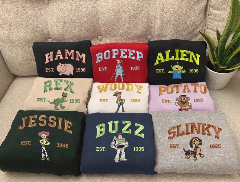 May include: A collection of embroidered sweatshirts featuring characters from the movie Toy Story. Each sweatshirt displays a character's name, the year 1995, and a corresponding image. Colors include black, red, green, white, and gray.