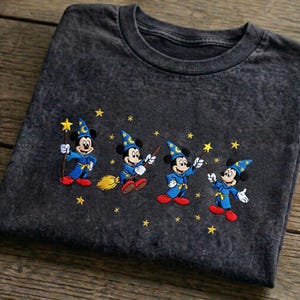 Könnte beinhalten: Dunkelgraues T-Shirt mit einem gestickten Design von Mickey Mouse als Zauberer. Mickey trägt einen blauen Umhang und Hut, hält einen Zauberstab oder Besen, mit gelben Sternen. Das T-Shirt ist auf einer Holzoberfläche gefaltet.