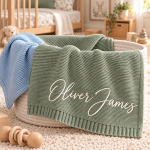 Könnte beinhalten: Eine salbeigrüne Strickdecke für Babys mit dem in weißer Schreibschrift eingestickten Namen Oliver James, drapiert über einem weißen Seilkorb neben einer hellblauen Strickdecke, mit einem strukturierten kraus rechts gestrickten Muster.