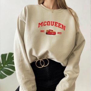 Puede incluir: Sudadera beige con la palabra "MCQUEEN" en rojo, un dibujo de coche rojo y el texto "EST. 1977". La sudadera se combina con pantalones negros y un cintur&oacute;n dorado.