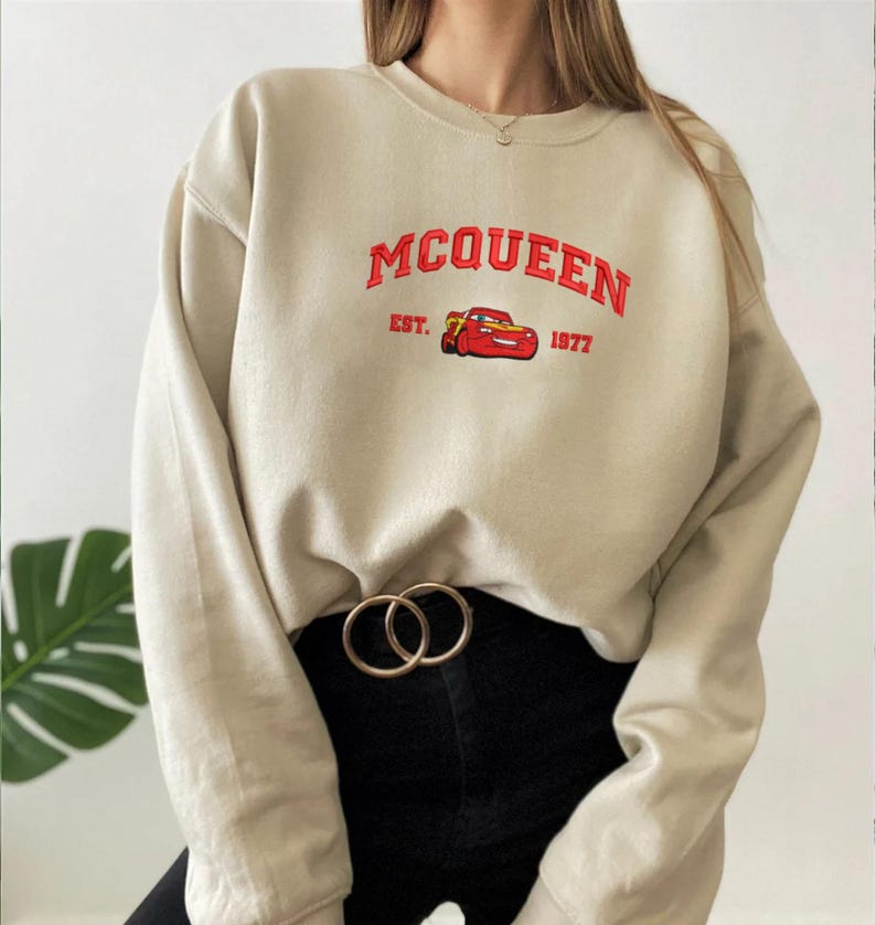 Puede incluir: Sudadera beige con la palabra "MCQUEEN" en rojo, un dibujo de coche rojo y el texto "EST. 1977". La sudadera se combina con pantalones negros y un cintur&oacute;n dorado.
