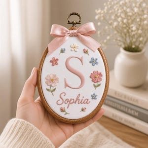 以下が含まれることがあります： 木製の楕円形刺繍枠に白い布が張られ、ピンクの糸で刺繍された文字「S」と筆記体の名前「Sophia」が施されています。ピンク、青、白の小さな花柄がデザインされており、上部にはピンクのサテンリボンが結ばれています。
