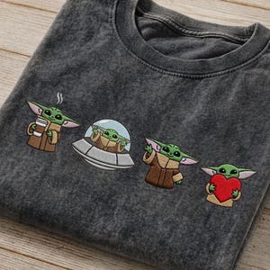 May include: Dark gray t-shirt featuring embroidered images of a green alien character in various poses. The alien is holding a cup, in a spaceship, waving, and holding a heart. The shirt is folded on a wooden surface.