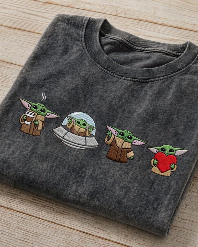 May include: Dark gray t-shirt featuring embroidered images of a green alien character in various poses. The alien is holding a cup, in a spaceship, waving, and holding a heart. The shirt is folded on a wooden surface.