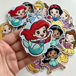 Op de afbeelding: Een verzameling geborduurde patches met cartoonprinsessen. De patches bevatten Ariel met rood haar en een zeegroene staart, Sneeuwwitje met een rode strik en Jasmine op een vliegend tapijt. Andere prinsessen zijn ook zichtbaar.