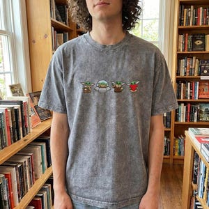 May include: A gray, short-sleeved t-shirt with a crew neck. The shirt features an embroidered design of four Grogu characters. The first Grogu is holding a cup, the second is in a spaceship, the third is holding a book, and the fourth is holding a heart.