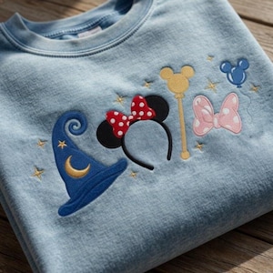 Pode incluir: Camisola azul clara com bordados tem&aacute;ticos da Disney. Apresenta um chap&eacute;u de feiticeiro azul com uma lua crescente dourada, orelhas da Minnie com um la&ccedil;o vermelho de bolinhas, uma varinha dourada, um la&ccedil;o rosa de bolinhas e uma cabe&ccedil;a do Mickey Mouse azul.
