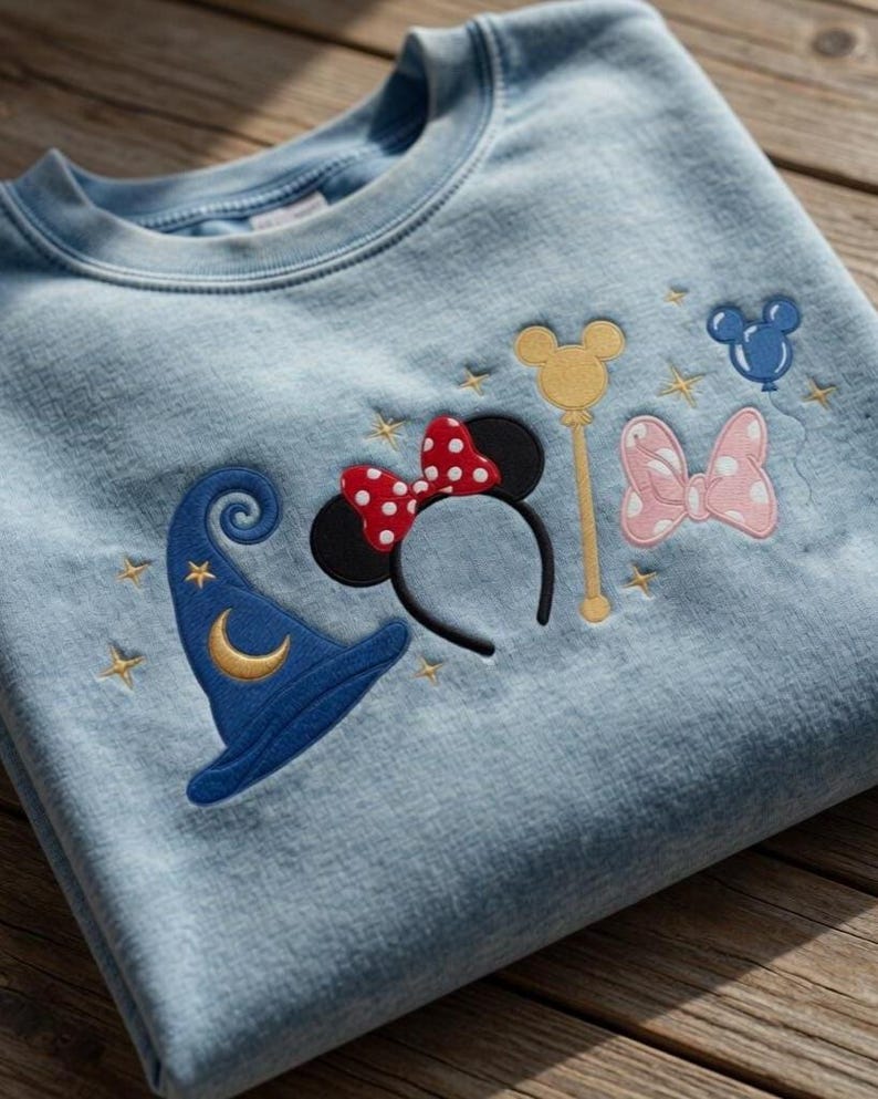 Pode incluir: Camisola azul clara com bordados tem&aacute;ticos da Disney. Apresenta um chap&eacute;u de feiticeiro azul com uma lua crescente dourada, orelhas da Minnie com um la&ccedil;o vermelho de bolinhas, uma varinha dourada, um la&ccedil;o rosa de bolinhas e uma cabe&ccedil;a do Mickey Mouse azul.