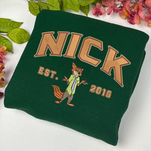 May include: A dark green sweatshirt with the name "NICK" embroidered in tan and orange. Below the name is "EST. 2016" and an embroidered cartoon fox character. The sweatshirt is folded and displayed on a white surface.