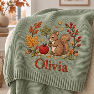 Könnte beinhalten: Eine salbeigrüne Strickdecke mit einem gestickten Motiv eines braunen Eichhörnchens, das eine Eichel hält, umgeben von bunten Herbstblättern, Beeren und einem roten Apfel. Der Name Olivia ist in orangefarbenem Text unter dem Eichhörnchen gestickt.