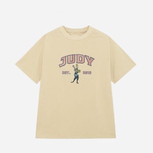 May include: A light beige t-shirt with the word "JUDY" in a pink and blue arched font. Below the word "JUDY" is the text "EST. 2016" and a cartoon rabbit character holding a camera. The t-shirt has short sleeves.