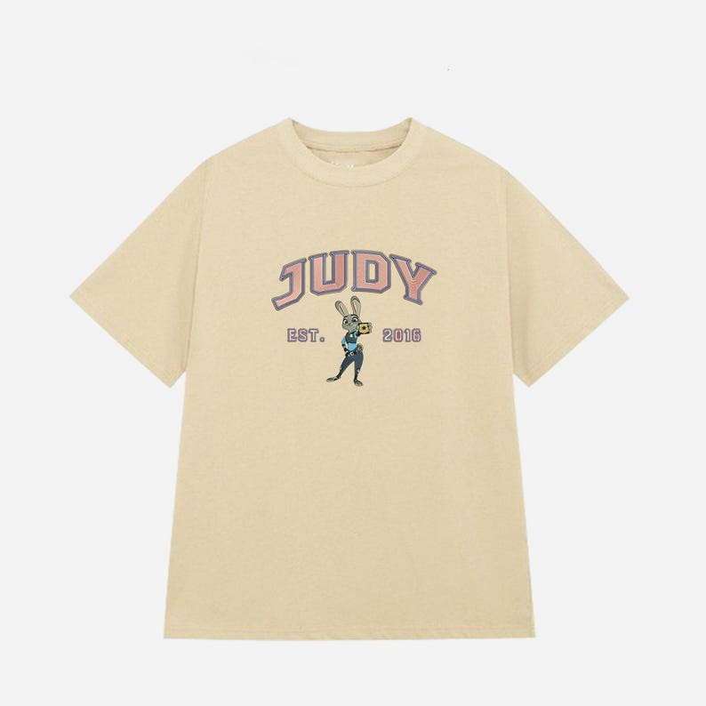 May include: A light beige t-shirt with the word "JUDY" in a pink and blue arched font. Below the word "JUDY" is the text "EST. 2016" and a cartoon rabbit character holding a camera. The t-shirt has short sleeves.
