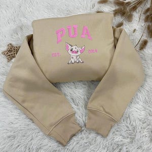 K&ouml;nnte beinhalten: Beigefarbener Pullover mit langen &Auml;rmeln und einem rosa gestickten Schwein mit dem Text "PUA EST. 2014" auf der Vorderseite.