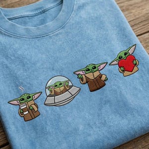 May include: Light blue crew neck sweatshirt with embroidered images of a green alien character in various poses. The alien is holding a coffee cup, in a spaceship, gesturing, and holding a red heart.