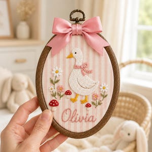 Op de afbeelding: Een ovale borduurring met een witte gans met een roze strik, omringd door geborduurde madeliefjes en rode paddenstoelen op roze en wit gestreepte stof. De naam Olivia is in roze draad geborduurd, bekroond met een roze satijnen strik.