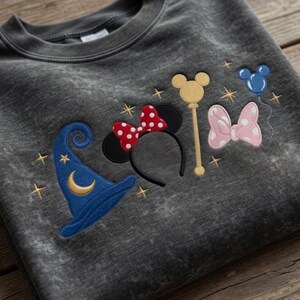 Pode incluir: Camisola cinza escuro com desenhos bordados com tema da Disney. Apresenta um chap&eacute;u de mago azul, orelhas da Minnie com um la&ccedil;o vermelho de bolinhas, uma varinha dourada, um la&ccedil;o rosa e um bal&atilde;o azul do Mickey Mouse. Estrelas douradas est&atilde;o espalhadas pelos desenhos.