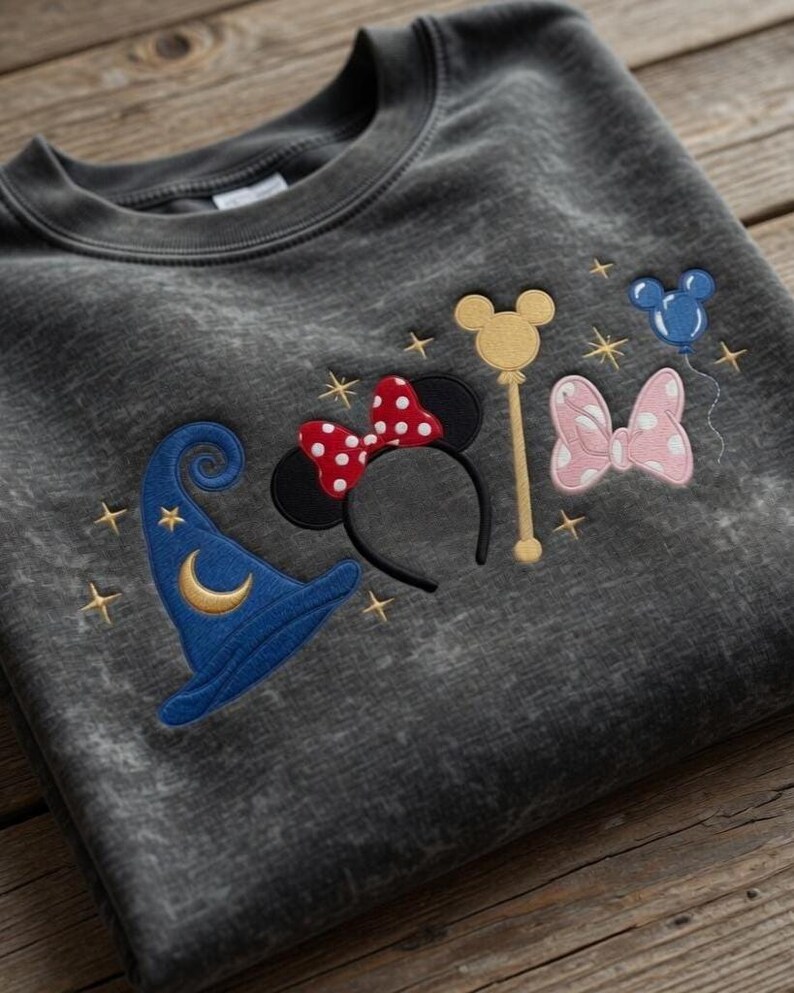 Pode incluir: Camisola cinza escuro com desenhos bordados com tema da Disney. Apresenta um chap&eacute;u de mago azul, orelhas da Minnie com um la&ccedil;o vermelho de bolinhas, uma varinha dourada, um la&ccedil;o rosa e um bal&atilde;o azul do Mickey Mouse. Estrelas douradas est&atilde;o espalhadas pelos desenhos.
