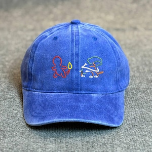 May include: A blue baseball cap with embroidered cartoon figures in red, green, and white. The cap has a curved brim and a button on top. The fabric appears to be a washed denim style.