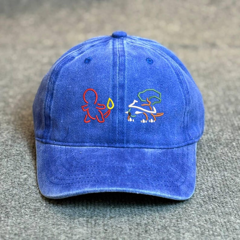 May include: A blue baseball cap with embroidered cartoon figures in red, green, and white. The cap has a curved brim and a button on top. The fabric appears to be a washed denim style.