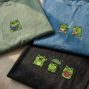 May include: Three t-shirts in sage green, blue, and black with embroidered frog designs. The green shirt features a frog reading a book. The blue shirt shows a frog with a lightsaber and another with a boba drink. The black shirt displays three frogs.