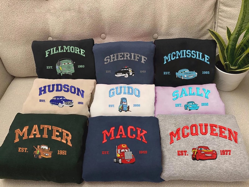 Puede incluir: Una colecci&oacute;n de sudaderas bordadas con personajes de la pel&iacute;cula Cars. Cada sudadera muestra el nombre de un personaje, un gr&aacute;fico de coche y el a&ntilde;o de establecimiento. Los colores incluyen negro, azul marino, verde, beige, blanco y gris.