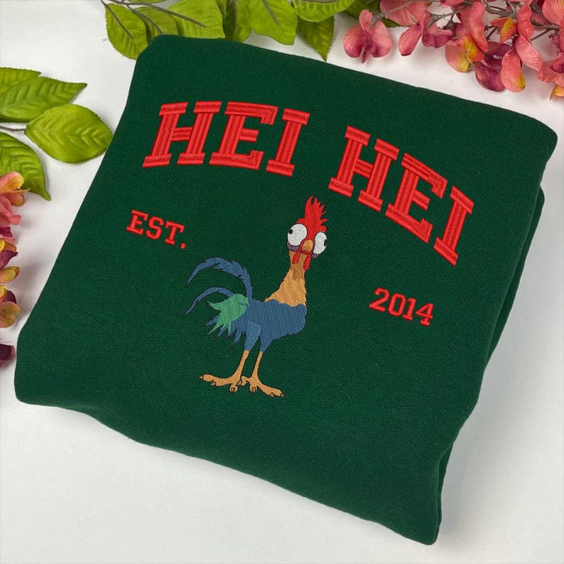 K&ouml;nnte beinhalten: Gr&uuml;ner Sweatshirt mit rotem gesticktem Text "Hei Hei" und den Worten "Est. 2014". Ein Cartoon-Hahn mit roten Augen und einem blau-gr&uuml;nen Schwanz ist auf der Vorderseite gestickt.