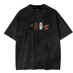 May include: Black acid wash t-shirt with a cartoon design of four characters: a black figure, a brown Wookiee, a white Stormtrooper, and a green alien holding a red heart. The design is across the chest.