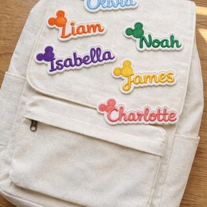 Pode incluir: Uma mochila creme com vários patches de nomes coloridos. Os patches apresentam nomes como Olivia, Liam, Isabella, Noah, James e Charlotte, cada um com um design inspirado no Mickey Mouse. A mochila tem um bolso frontal e um zíper.