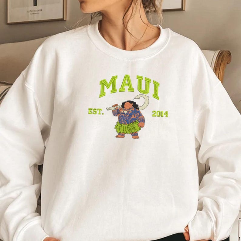 K&ouml;nnte beinhalten: Wei&szlig;er Pullover mit einem gr&uuml;nen Aufdruck von Maui aus dem Film Vaiana. Der Aufdruck enth&auml;lt den Text "MAUI" in einem gr&uuml;nen Bogen, "EST. 2014" unter dem Bogen und ein Cartoon-Bild von Maui, der seinen Angelhaken h&auml;lt.