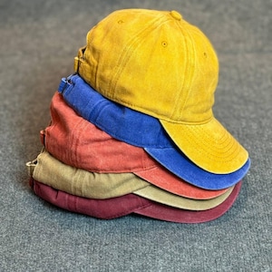 May include: A stack of six baseball caps in various colors: yellow, blue, red, tan, and burgundy. The caps have a worn, vintage look, with curved visors and adjustable straps.