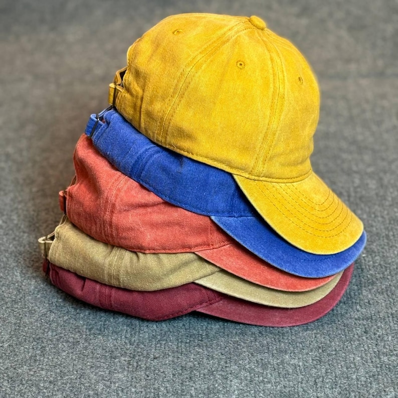 May include: A stack of six baseball caps in various colors: yellow, blue, red, tan, and burgundy. The caps have a worn, vintage look, with curved visors and adjustable straps.