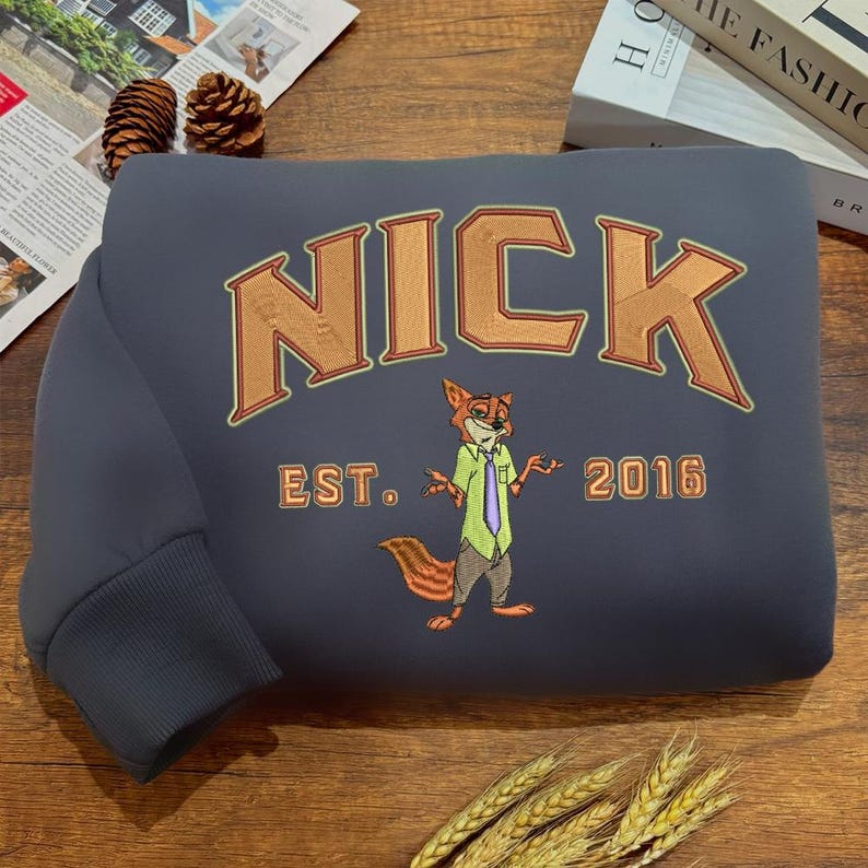 May include: A dark gray sweatshirt with the name "NICK" in orange block letters. Below the name is an embroidered image of a fox in a shirt and tie, with the text "EST. 2016". The sweatshirt has a ribbed cuff.