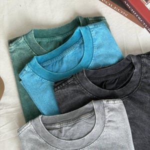 May include: A stack of five t-shirts in various colors: green, blue, black, and gray. The shirts have a vintage, washed-out appearance. The collars are ribbed and the fabric appears soft.