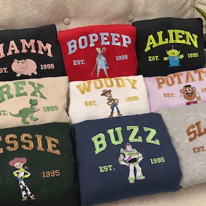 May include: A collection of embroidered sweatshirts featuring characters from a popular animated film. Each sweatshirt is a different color and features a character's name, a graphic of the character, and the year "1995".