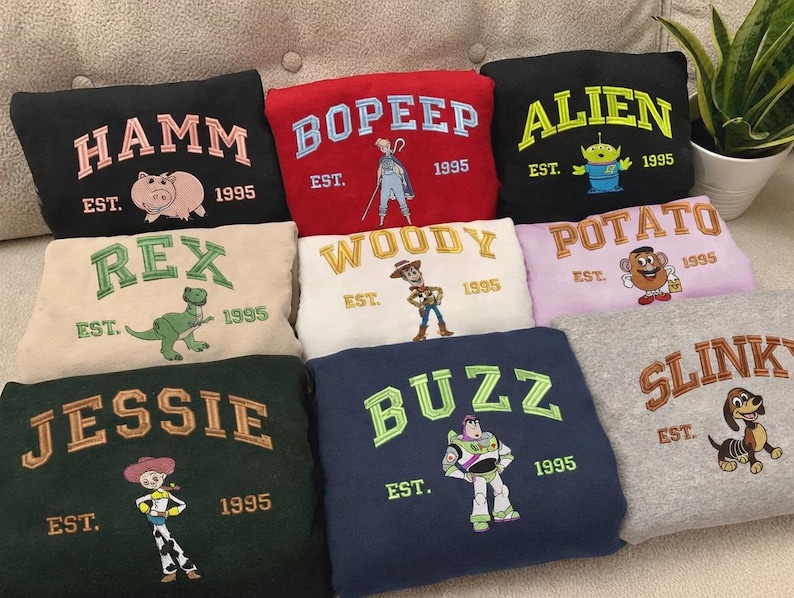 May include: A collection of embroidered sweatshirts featuring characters from a popular animated film. Each sweatshirt is a different color and features a character's name, a graphic of the character, and the year "1995".